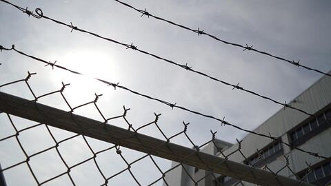 Maschendrahtzaun Maschendraht- und Stacheldrahtzaun, im Hintergrund ein Teil eines Betongebäudes.